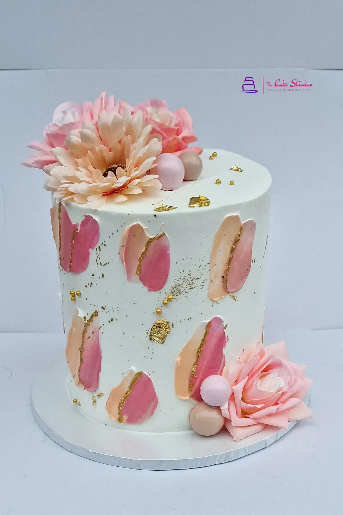 A pink and gold anniversary cake with palette-knife brushstrokes, gold-leaf flecks, and fresh peach blooms, baked by The Cake Studio Harare