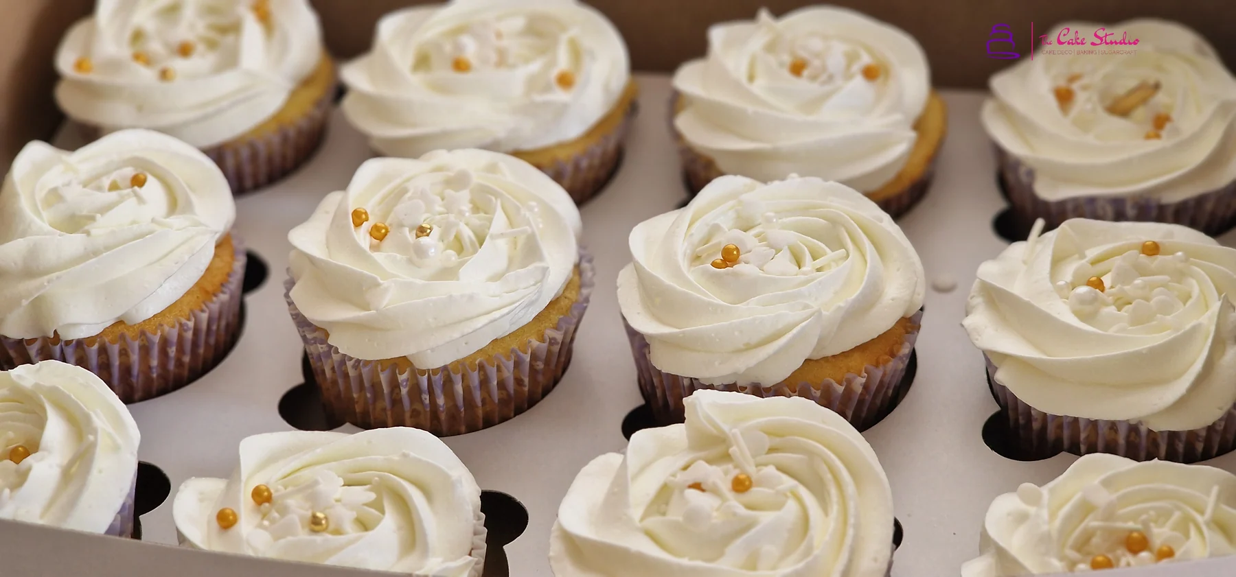 A 12-piece box of ivory vanilla buttercream rose-piped cupcakes scattered with gold sugar pearls, by The Cake Studio Harare