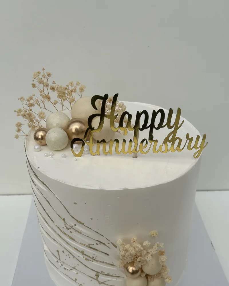 A white buttercream anniversary cake with combed texture, gold brushwork, cream and gold spheres, dried baby’s breath and a gold Happy Anniversary topper, by The Cake Studio Harare