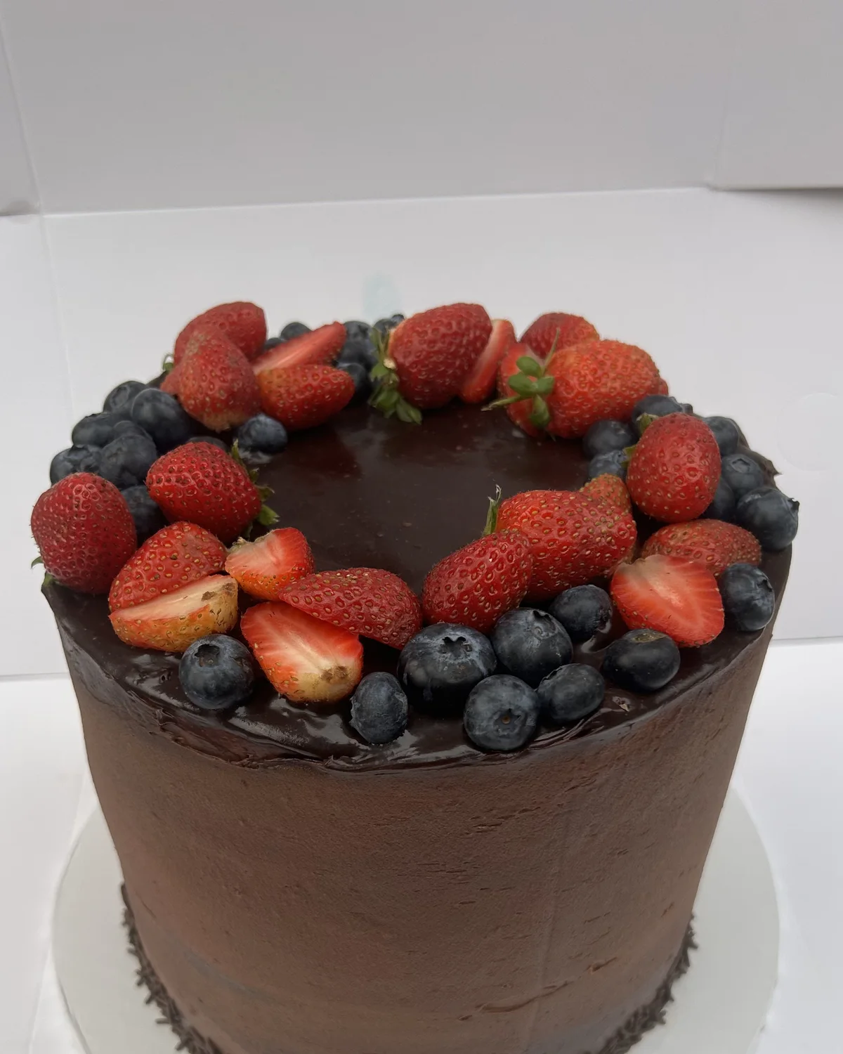 An ultra moist chocolate cake covered in dark chocolate ganache, ringed with strawberries and blueberries with chocolate sprinkles at the base, by The Cake Studio Harare