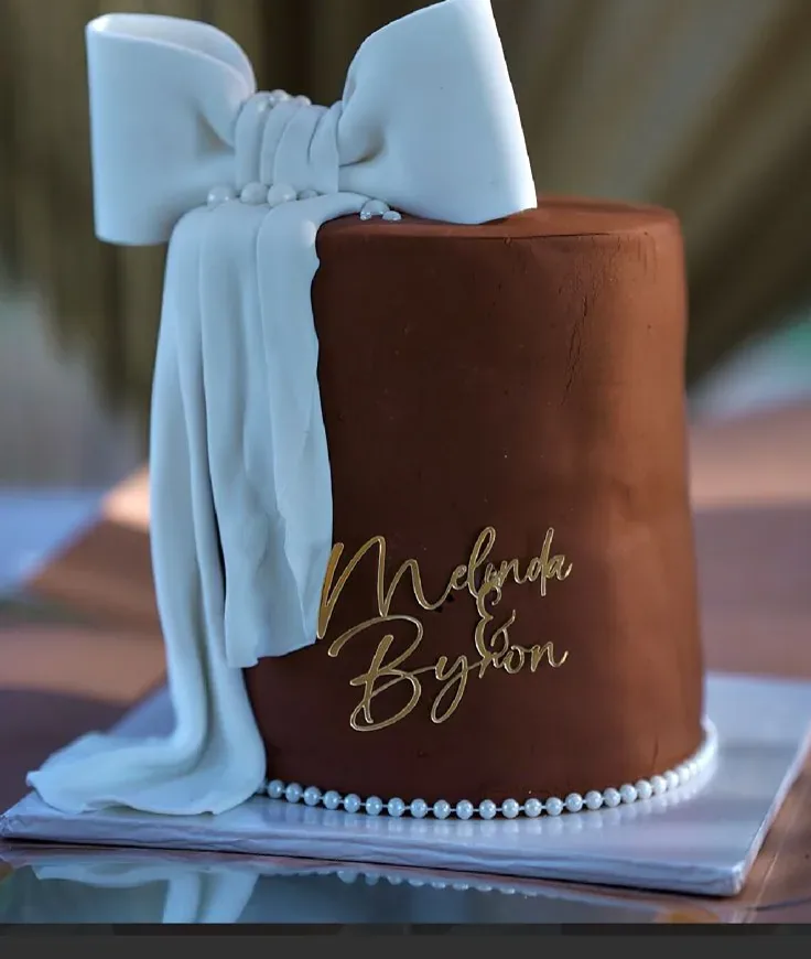 A single-tier chocolate-finished lobola cake with a fondant bow and gold names, by The Cake Studio Harare