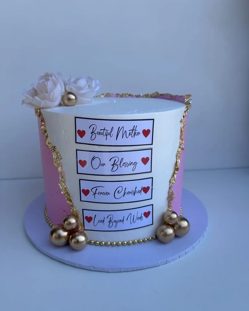 A pink and white buttercream cake split by a torn seam of gold leaf, with gold spheres, cream roses and four printed plaques reading Beautiful Mother, Our Blessing, Forever Cherished and Loved Beyond Words, by The Cake Studio Harare