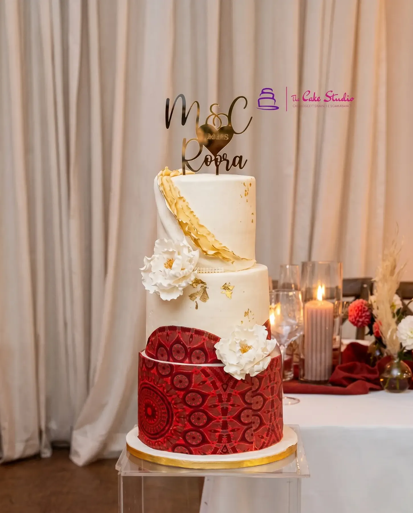 A three-tier Roora Day cake by The Cake Studio Harare with sugar peonies, gold leaf, an African-pattern red bottom tier, and a gold ‘M&C Roora’ topper